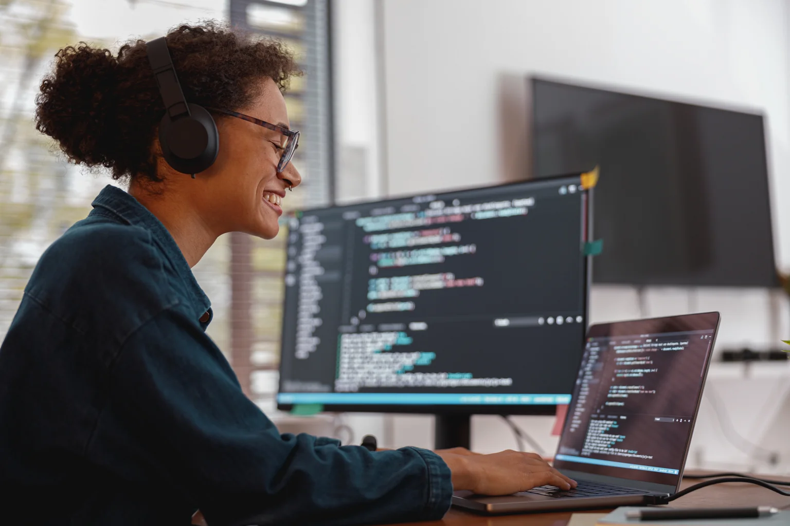 Mulher de óculos e fones trabalhando no notebook, com código aberto em um monitor ao lado.